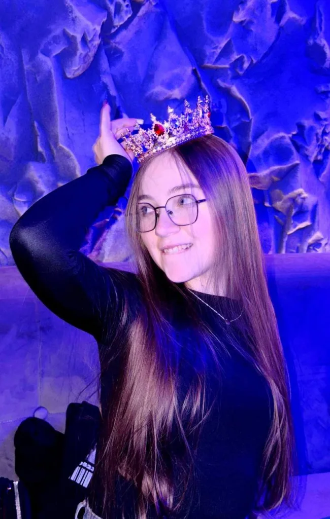 ArrinaWairr smiles while wearing glasses and a gold crown, with her long brown hair down against a blue background.