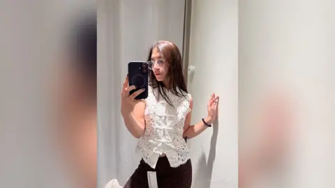 Young woman in a white lace sleeveless top taking a mirror selfie in a bright bathroom.