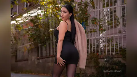 Elegant woman in a black mini dress and tights posing outdoors near a garden fence in soft natural light.