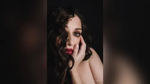 Moody close-up portrait of a woman with wavy dark hair and dramatic smoky eye makeup against a black background.