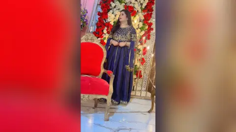 Elegant woman in navy blue sequined traditional Indian outfit standing before a red floral backdrop.