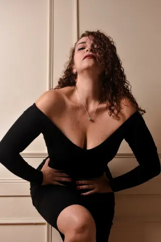 Elegant woman in a black off-the-shoulder dress posing confidently against a neutral wall.
