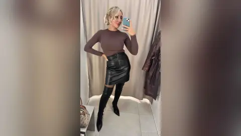 Blonde woman in a brown long-sleeve top and black leather mini skirt posing for a mirror selfie in a bright room.