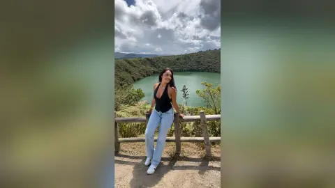 FabianaLima stands behind a wooden fence wearing a black tank top and light blue jeans against a backdrop of a green lake and mountains.