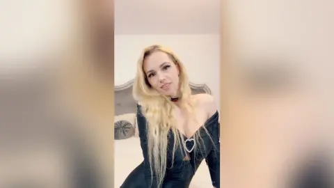 Smiling blonde woman in a black off-the-shoulder dress posing in a bright bedroom setting.