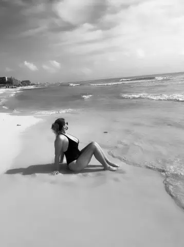 Woman in black one-piece swimsuit sitting on a sunny beach by the ocean waves, black and white aesthetic.