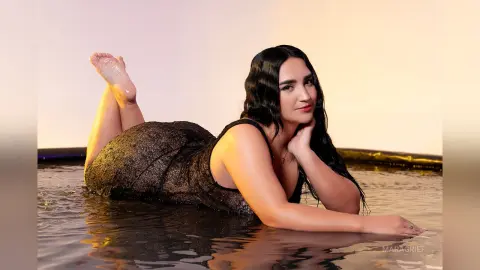 MaraGrief poses lying on her stomach in a pool of water, wearing a black sparkly dress with long dark hair.
