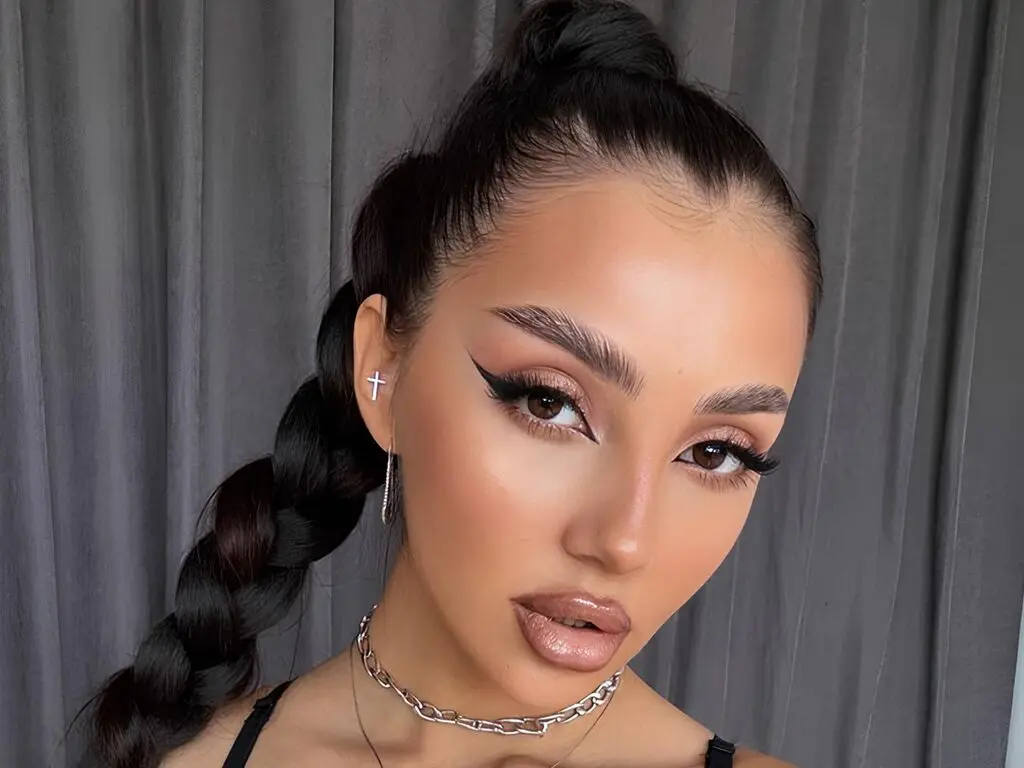 Woman with sleek braided hairstyle and glamorous winged eyeliner makeup posing against a grey curtain background.