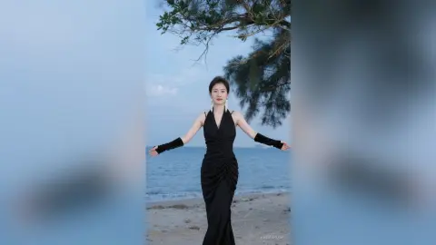 Elegant woman in a black evening gown and gloves posing on a serene beach under a leafy tree.