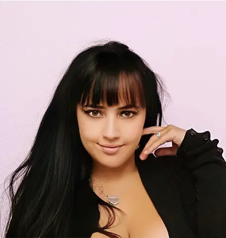 Smiling brunette woman with bangs wearing a black cardigan and heart necklace against a pink background.