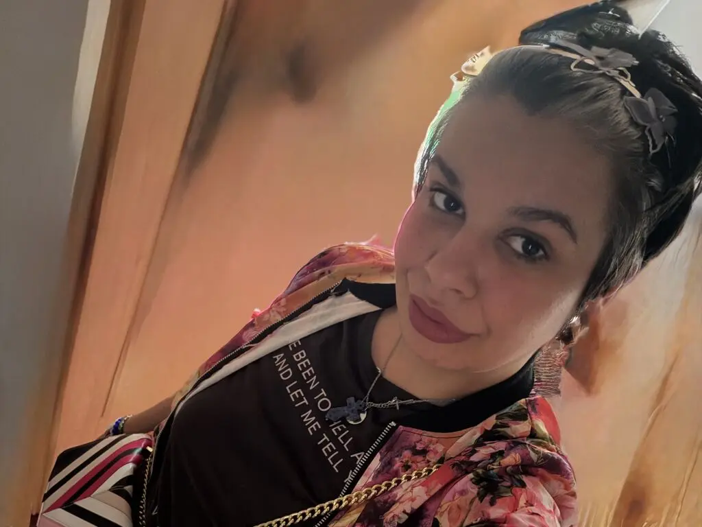 SabrinFisher looks at the camera wearing a black t-shirt with white text and a floral print jacket, her dark hair pulled back.