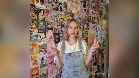 LisetteMaupredi poses in denim overalls and a white t-shirt, flashing peace signs in front of a wall covered in anime posters.