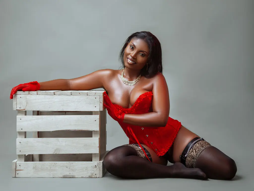 Smiling woman in red lace lingerie and black stockings sitting by a white wooden crate in a studio setting.
