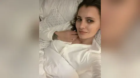 Smiling woman in white pajamas relaxing on a bed in a cozy, bright bedroom setting.