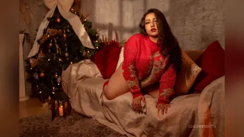 Curvy woman in a red festive sweater sitting on a cozy sofa near a glowing Christmas tree in a warm holiday setting.