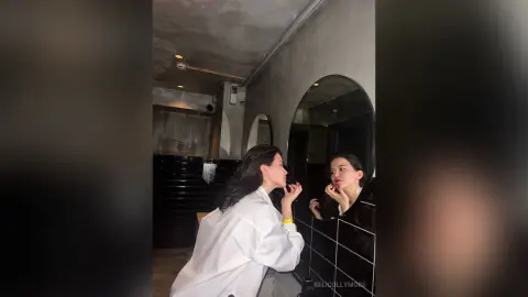 Young woman in a white oversized shirt applying lipstick in a moody, modern dark-toned restroom mirror.