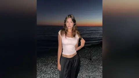 JazminHalajian poses on a beach at dusk wearing a tight white tank top and black shorts, with the ocean and sunset in the background.
