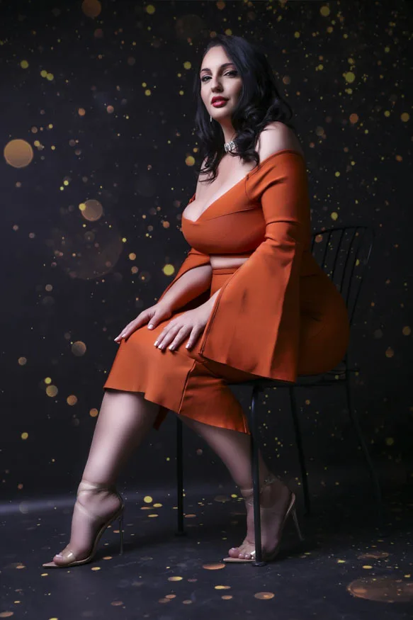 JudithConner poses in a black bodysuit and black fishnet tights, showing her cleavage and dark hair against a warm, indoor background.
