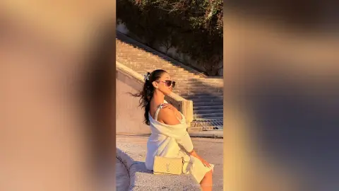 Stylish woman in a white off-the-shoulder dress and sunglasses sitting on stone stairs in warm sunlight.
