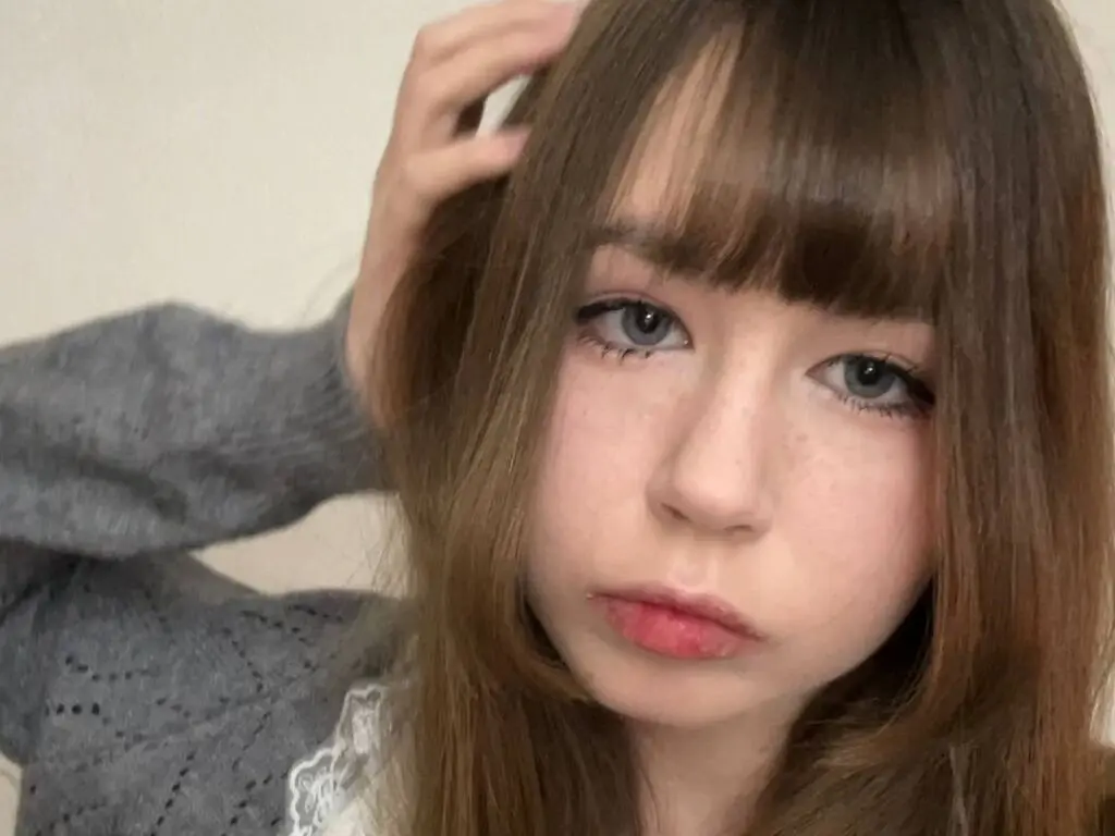 Young woman with brown bangs and freckles wearing a grey sweater in a soft, natural indoor setting.