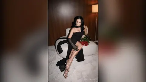 Elegant woman in a black evening gown and gloves sitting on a white chair holding red roses in a moody setting.