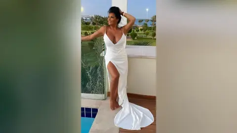 MissyJolie poses on a balcony wearing a white low-cut slit dress with her hand in her hair.