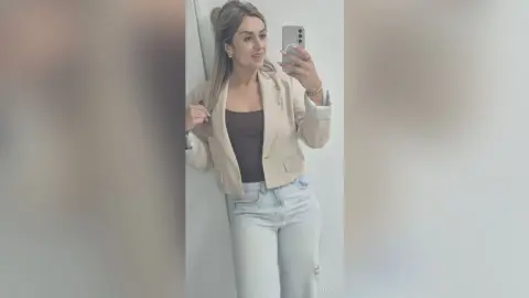 Jess poses in a light tan blazer and dark tank top with light wash jeans, holding her phone for a mirror selfie.