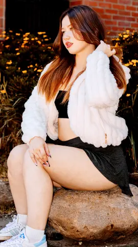Young woman in a white fuzzy jacket and black crop top sitting outdoors on a rock near garden flowers.