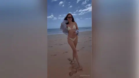 Stylish woman in a white bikini and sunglasses posing on a sunny beach under a blue sky.