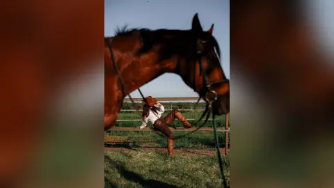 Cowgirl in western attire sitting in a grassy field next to a large brown horse under a clear sky.