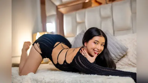 Smiling woman in a black ribbed knit cutout bodysuit relaxing on a white bed in a cozy bedroom setting.