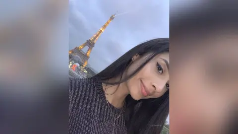 LucyMartin smiles while posing outdoors with the Eiffel Tower visible in the background behind her long dark hair.