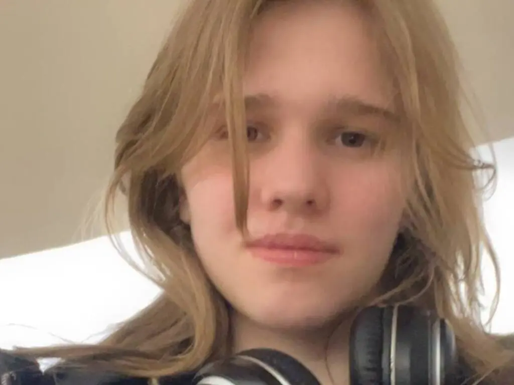 Young woman with long blonde hair wearing black headphones in a bright, soft-lit indoor setting.