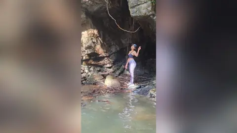 MaddyBrohms poses in a dark cave setting, wearing a black bra and tight grey leggings while standing near a small pool of water.
