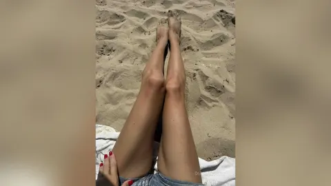 DianKrouse — DianKrouse lies on a white towel on the sand with her legs stretched out in denim shorts.