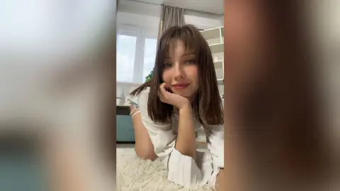 Smiling young woman with brown hair in a white shirt relaxing on a fluffy white rug in a bright room.