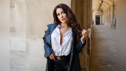 Stylish woman in a white blouse and black leather jacket posing in a sunlit stone hallway.
