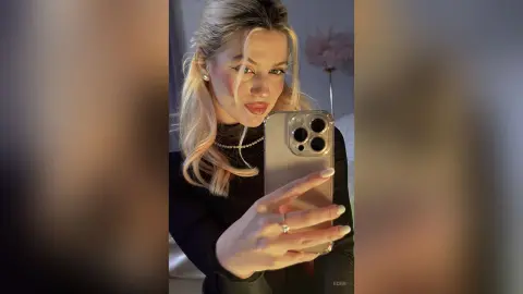 Blonde woman in a black turtleneck and gold necklace taking a glamorous selfie in a soft-lit bedroom.