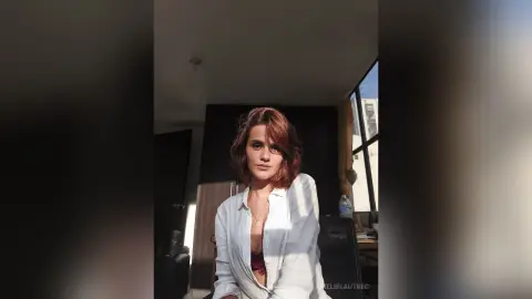 AmelieLautrec poses in a white button-down shirt that is open to show her red lace bra, with her reddish-brown hair styled down in a room with bright sunlight.