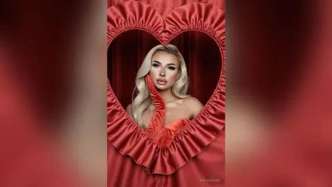 Glamorous blonde woman in red velvet gloves posing inside a heart-shaped red curtain frame.