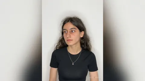FondaYarbro — FondaYarbro wears a black t-shirt and a silver necklace while looking to the side against a white wall.