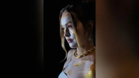AliceTucci looks at the camera with gold paint or liquid decorating her face, neck, and chest against a dark background.