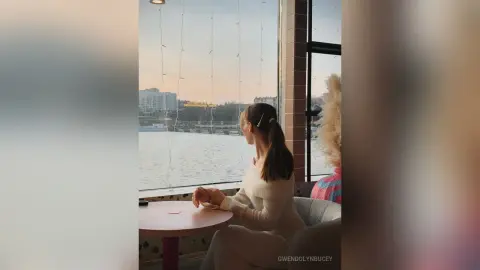 GwendolynBucey sits by a large window overlooking a lake, wearing a white long-sleeved top and looking out at the water.