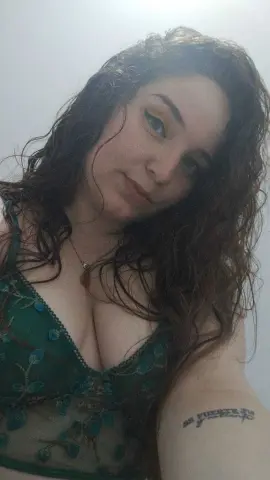 Young woman with long wavy hair wearing a green floral lace camisole in a soft, natural light setting.
