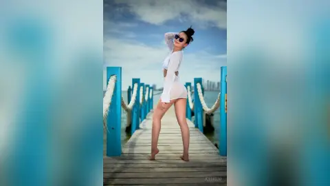JessieLiu stands on a wooden pier wearing a white long-sleeved bodysuit and sunglasses against a blue sky.
