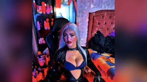 Woman with long silver hair in a black leather outfit sitting in a bedroom with moody red lighting.