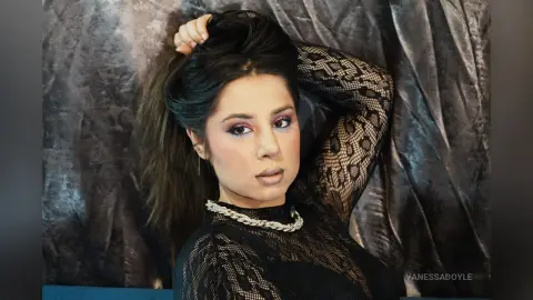Glamorous woman in a black lace long-sleeve top posing against a textured grey backdrop with moody lighting.