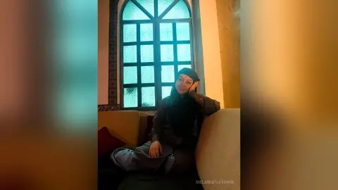 Smiling woman in a black hijab and dark outfit sitting on a sofa by a window in a warm, cozy room.