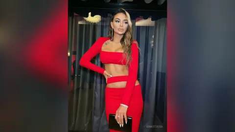 Anastasia poses in a red long-sleeved crop top and matching red skirt, holding a black clutch bag.