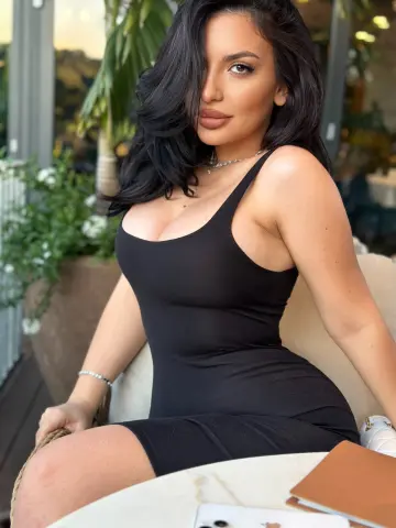 NoraSin poses in a tight black tank dress with her long dark hair down, sitting at a white table outdoors.